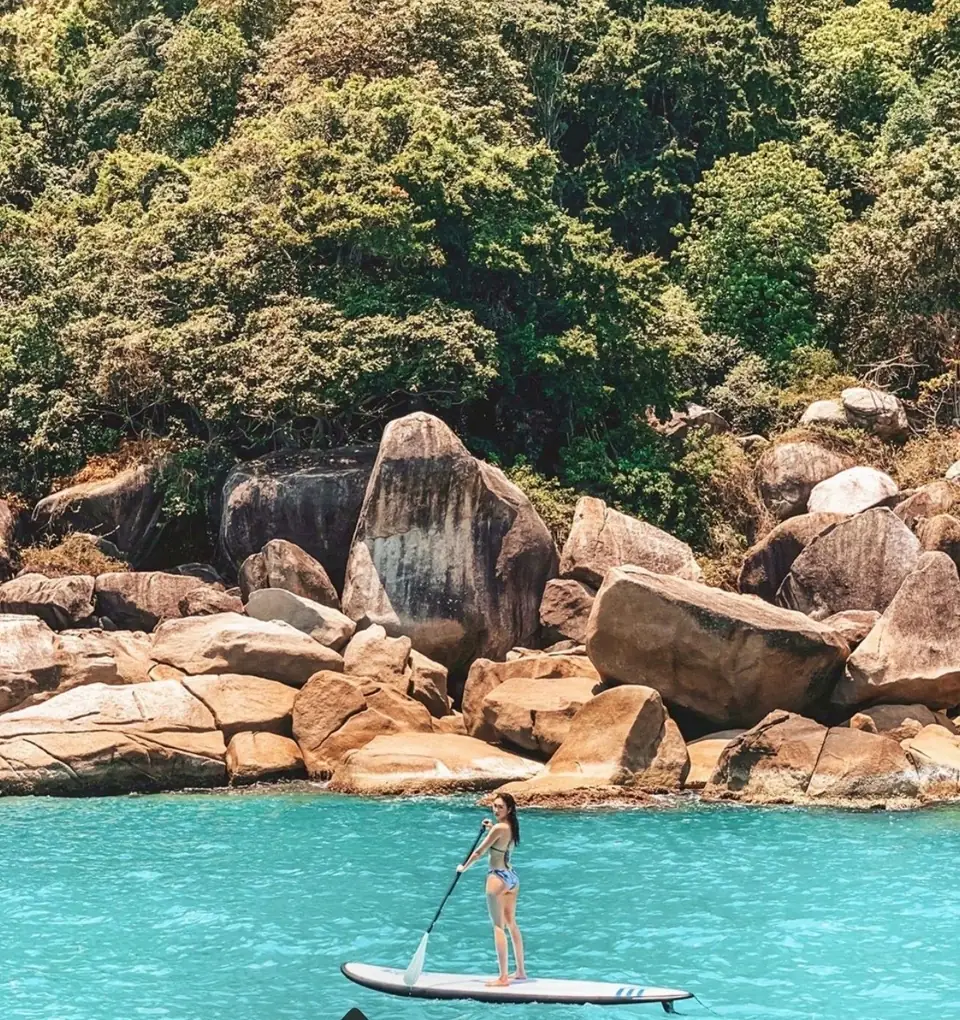A guest paddleboarding beside a rocky, tree-lined shoreline